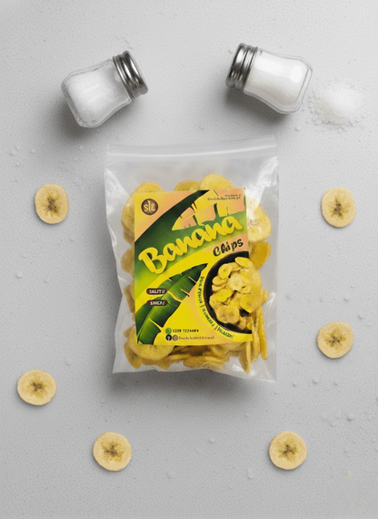 Packaged salted banana chips with a salt shaker on a grey surface with green leaf accents.